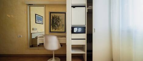 Une pièce avec un plancher en bois, une chaise blanche, un bureau avec un ordinateur, une armoire blanche et un miroir qui reflète un lit et un tableau accroché au mur.