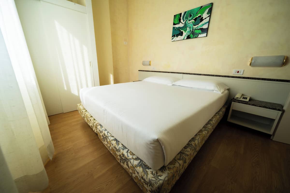 A neatly made bed in a room with a wooden floor, a white door, and a small bedside table with a lamp.