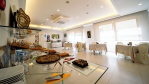 A spacious room with a buffet table displaying various desserts, white tables with chairs, and large windows allowing natural light. - Hotel Rosabianca (Rimini)