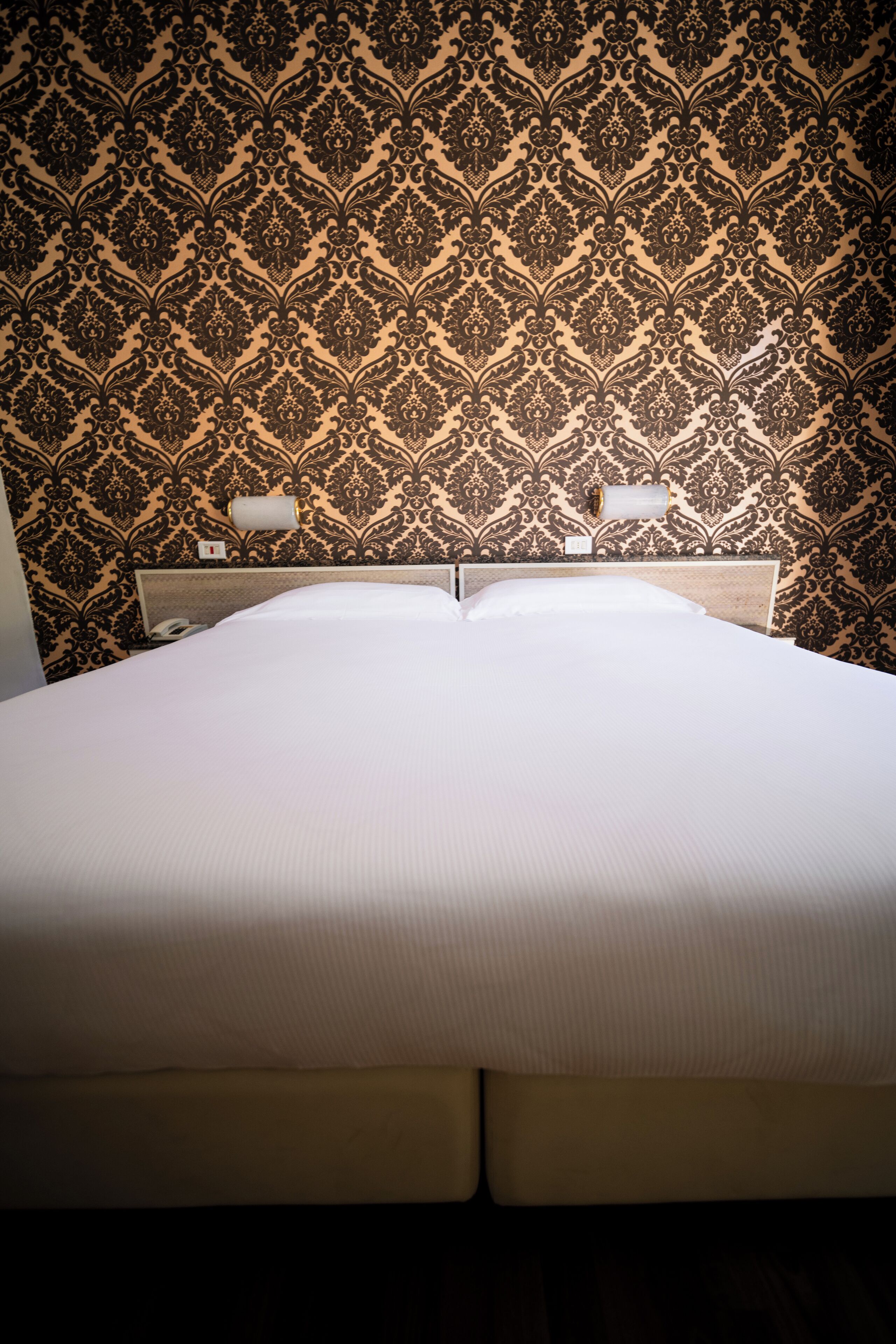 A hotel room with a large bed, a headboard with a light fixture, and a patterned wallpaper.