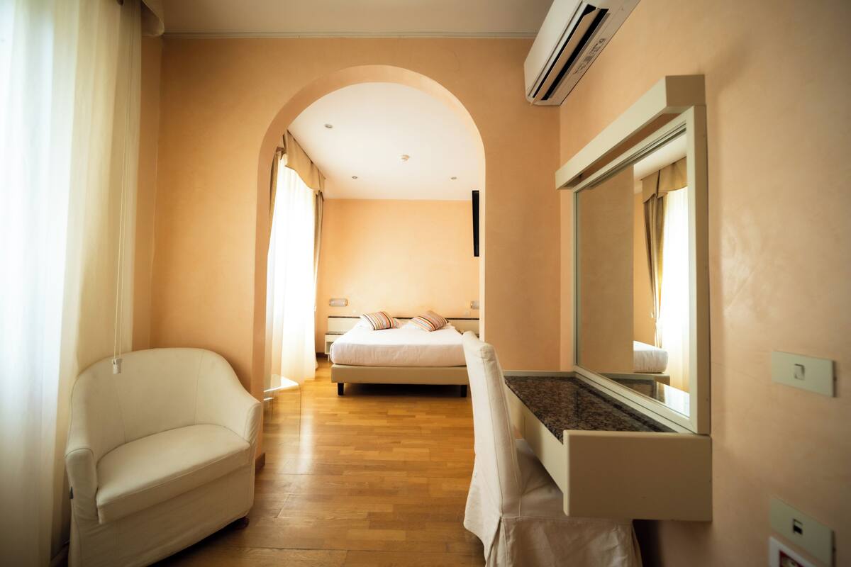 A hotel room with a bed, a chair, a built-in TV, and a large mirror.