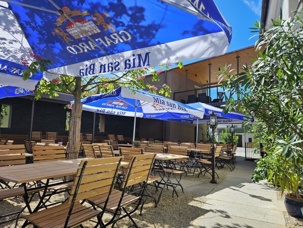 Outdoor seating with tables and chairs, umbrellas, and a building with a blue and white color scheme. - Stieglers - Restaurant Hotel Biergarten (Ergoldsbach)