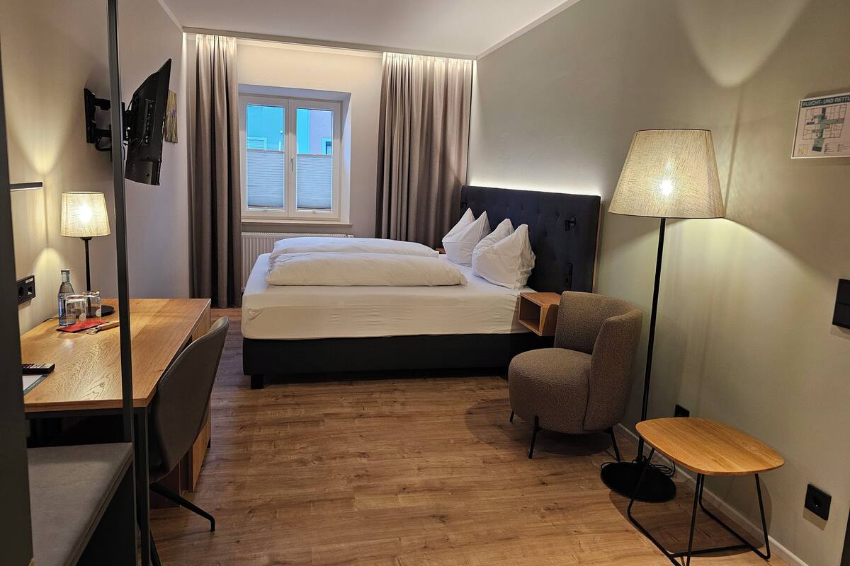 Une chambre d’hôtel avec un lit, un bureau, une chaise, une lampe et une fenêtre avec des rideaux.