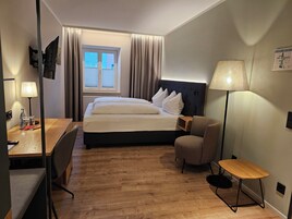 Quarto de hotel com cama, escrivaninha, cadeira, abajur e janela com cortinas.