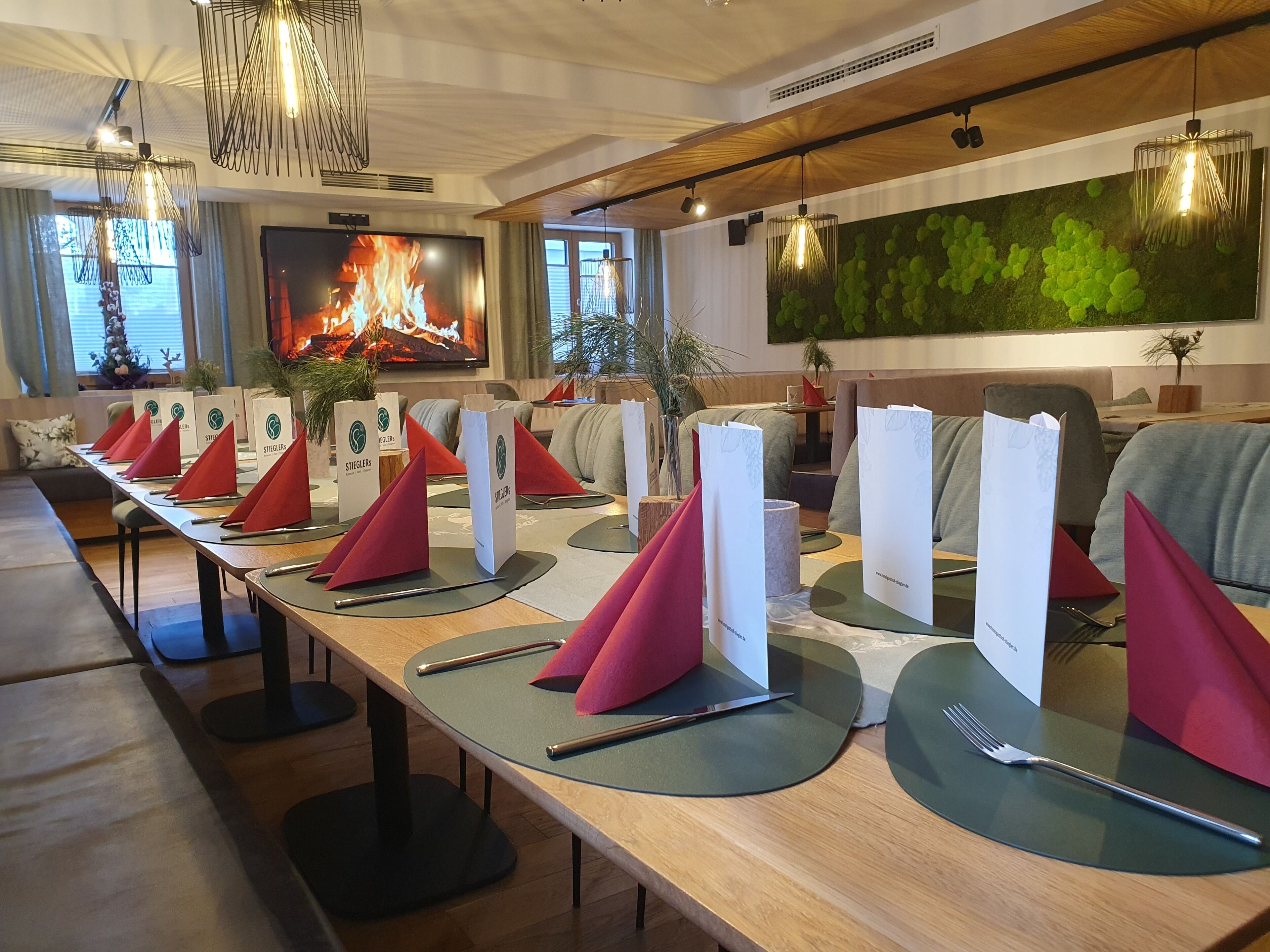 Una larga mesa de comedor dispuesta con servilletas rojas, menús blancos y cubiertos, con una chimenea y decoración de pared verde al fondo.