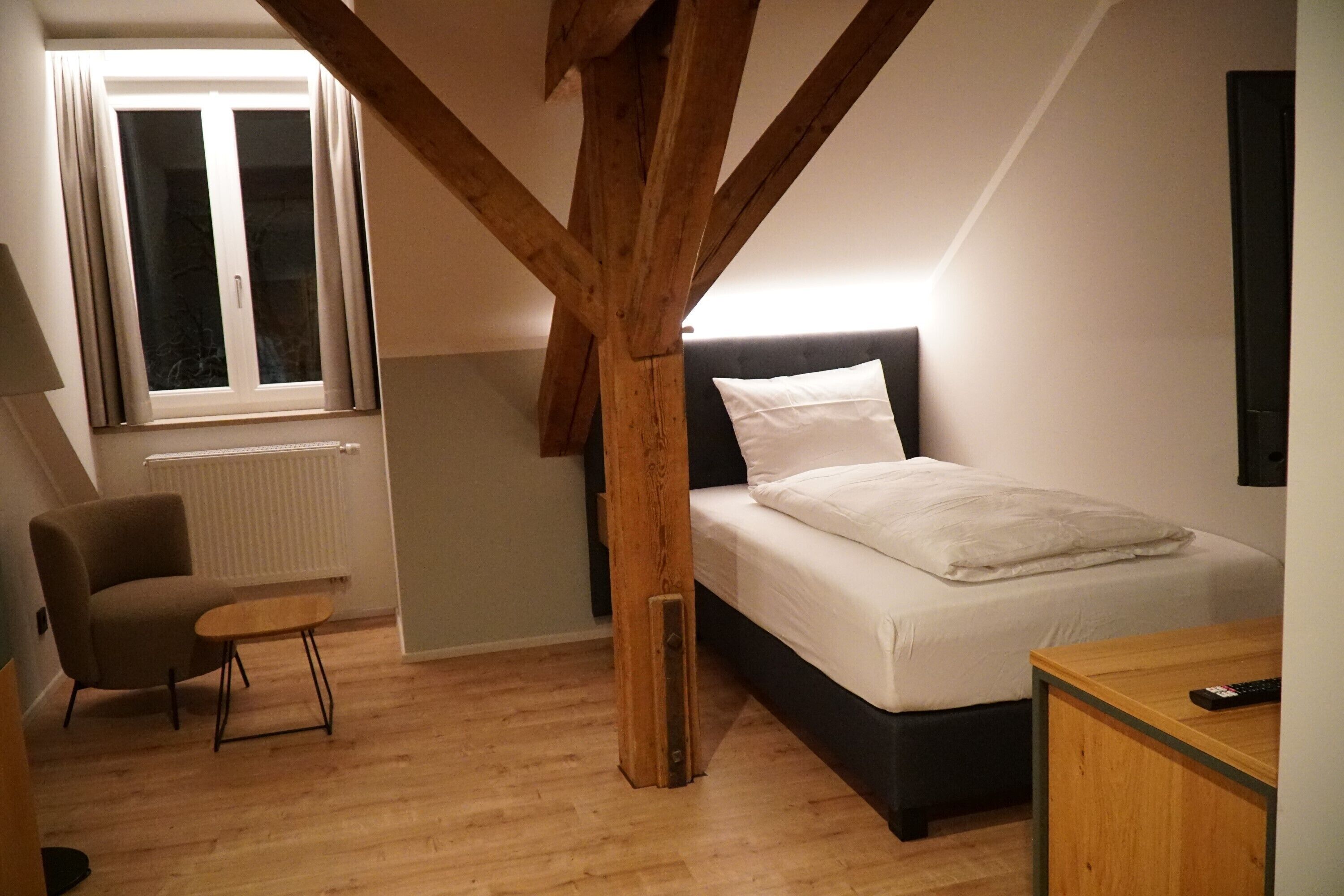 Un dormitorio con cama, una silla, una mesita, un ventanal y vigas de madera.