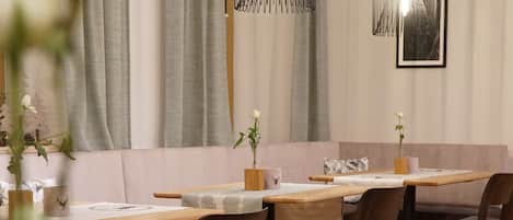 Un restaurante con mesas y sillas de madera, cortinas grises y lámparas colgantes.