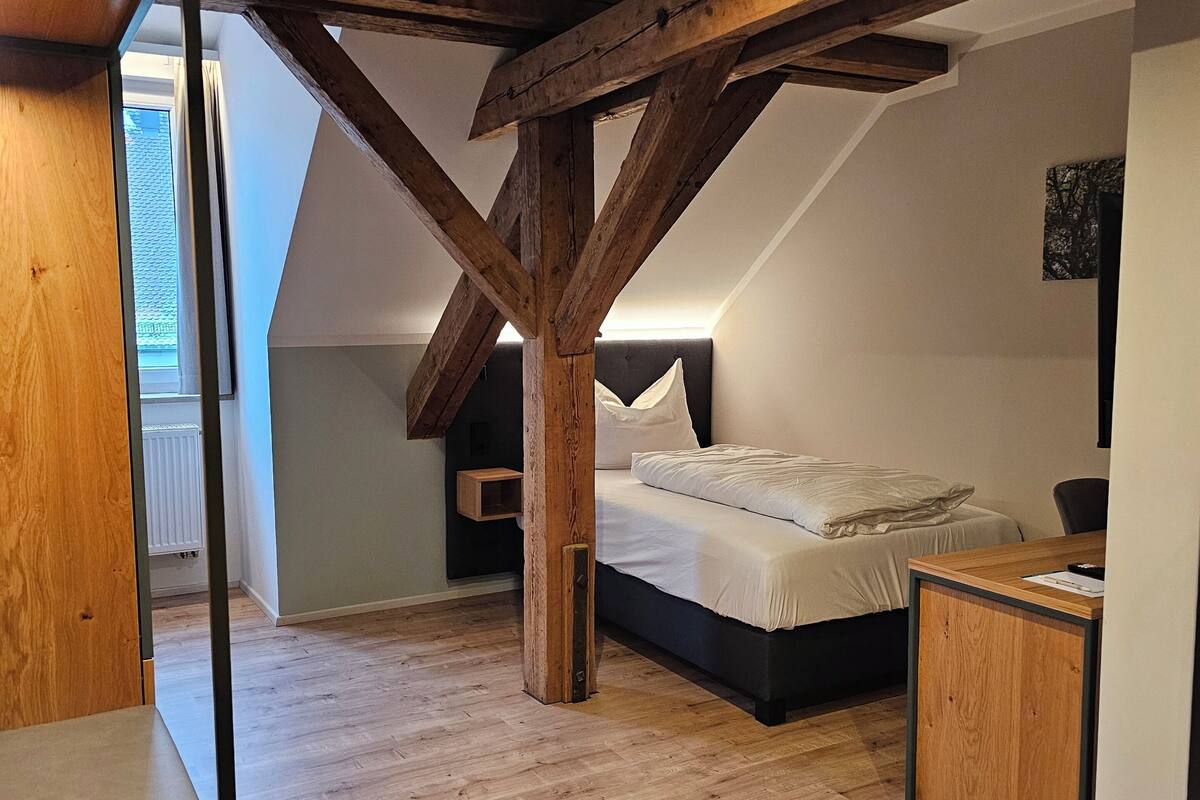 Une chambre avec un plafond en pente, des poutres en bois, un lit, un bureau et une fenêtre.