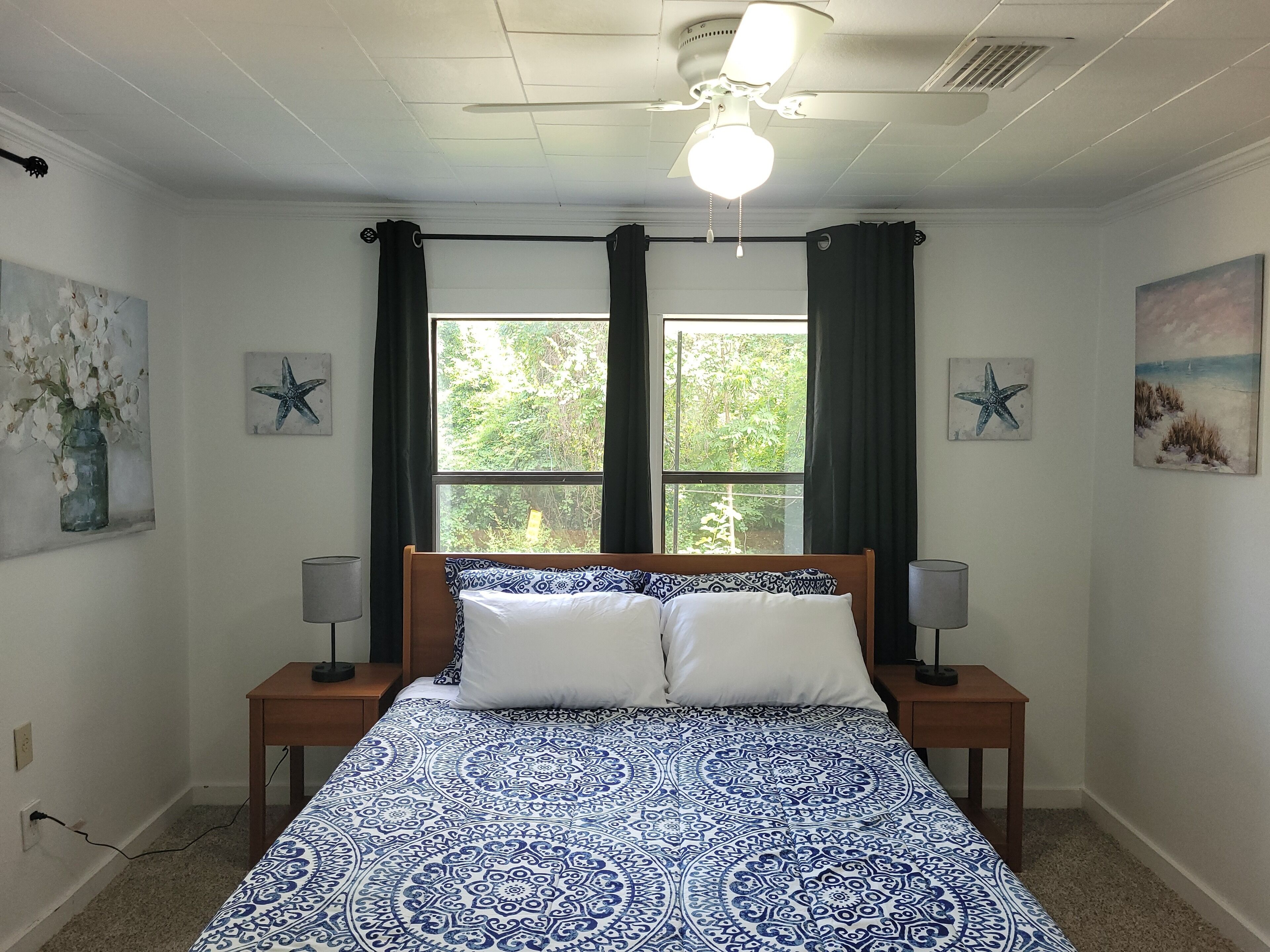 A bedroom with a bed, two bedside tables, a ceiling fan, a window with curtains, and wall art.
