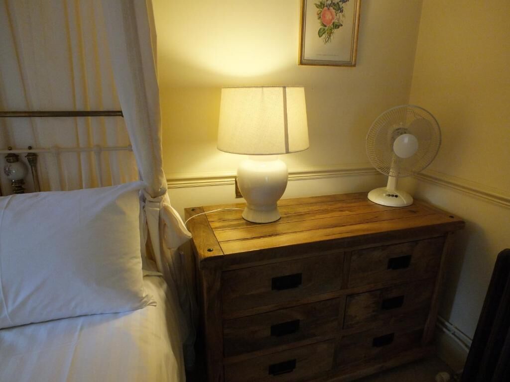 A bedroom with a bed, a bedside table with a lamp and a fan, and a wooden chest of drawers.