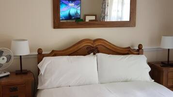 A neatly made bed with white linens, a wooden headboard, and two bedside lamps. A television is mounted on the wall above the bed, and a mirror is also visible.
