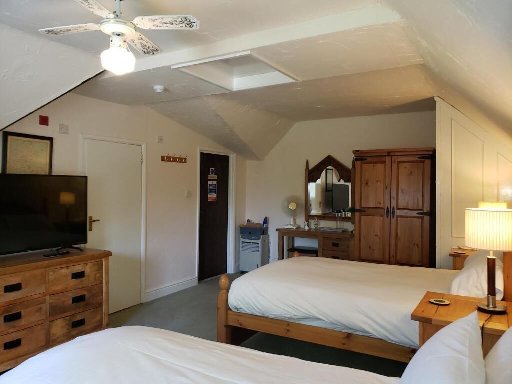 A hotel room with two beds, a wooden dresser, a television, a desk with a lamp, and a ceiling fan.