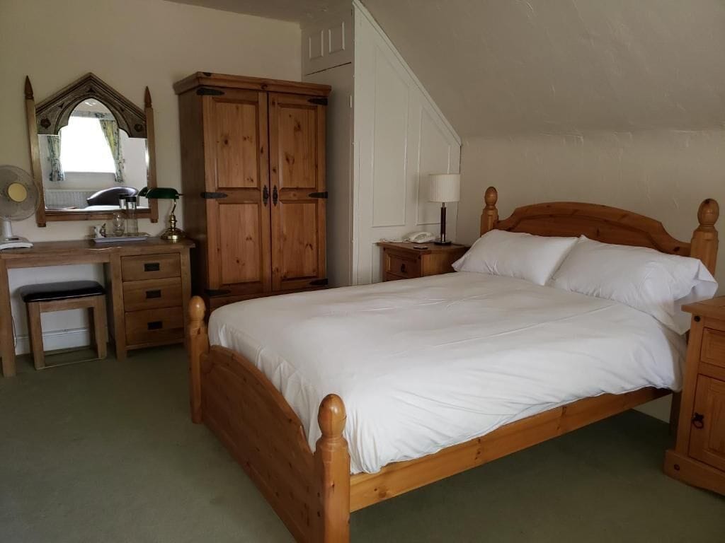 A bedroom with a wooden bed, a wooden wardrobe, a bedside table with a lamp, and a desk with a mirror and lamp.