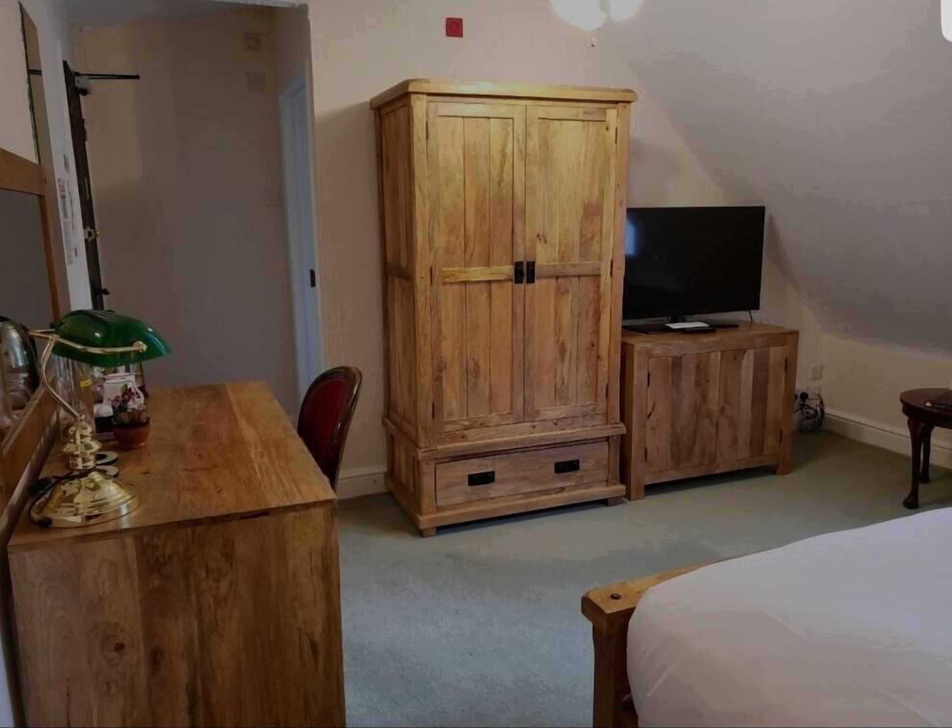 A room with a wooden wardrobe, a television on a small cabinet, a desk with a lamp, and a bed.