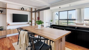Un salon moderne avec une table à manger en bois, des chaises noires, un téléviseur à écran plat fixé au mur et une grande fenêtre donnant sur la ville.