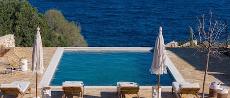 A rectangular pool with a stone border, surrounded by wooden lounge chairs and two white umbrellas.