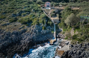 A coastal property with a stone bridge leading to a pink villa surrounded by lush greenery and rocky terrain. - Evilia Beach Villa With Private Pool (Zakynthos)