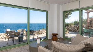 A modern living room with a large sliding glass door leading to an outdoor patio with a stone wall and chairs. - Evilia Beach Villa With Private Pool (Zakynthos)