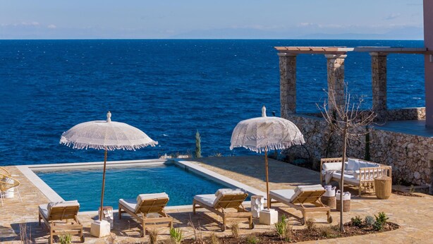 A poolside area with two white crocheted umbrellas, several wooden lounge chairs, and a stone pillar structure. - Evilia Beach Villa With Private Pool (Zakynthos)