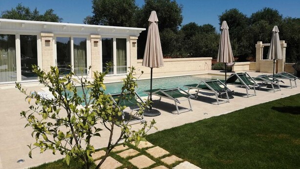 A pool area with lounge chairs and umbrellas, a covered seating area, and a building with large windows. - Villa Adriatica Near Ostuni Puglia (Ostuni)