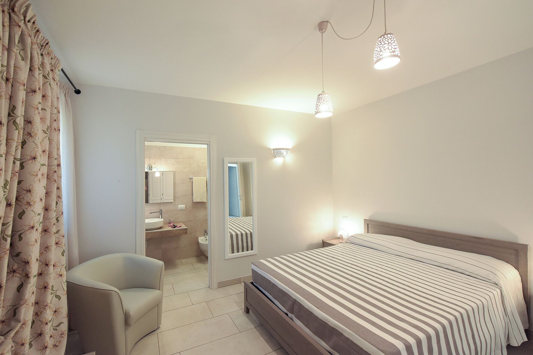 A bedroom with a bed, a chair, a nightstand, a mirror, and a bathroom visible through an open door.