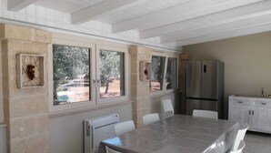 A dining area with a table, chairs, a refrigerator, and a window with a view of trees. - Villa Adriatica Near Ostuni Puglia (Ostuni)