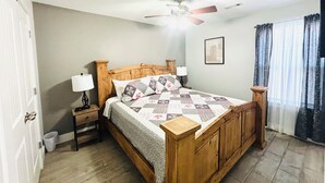 Une chambre à coucher avec un lit en bois, des tables de chevet, un ventilateur de plafond, une fenêtre avec des rideaux et une porte.