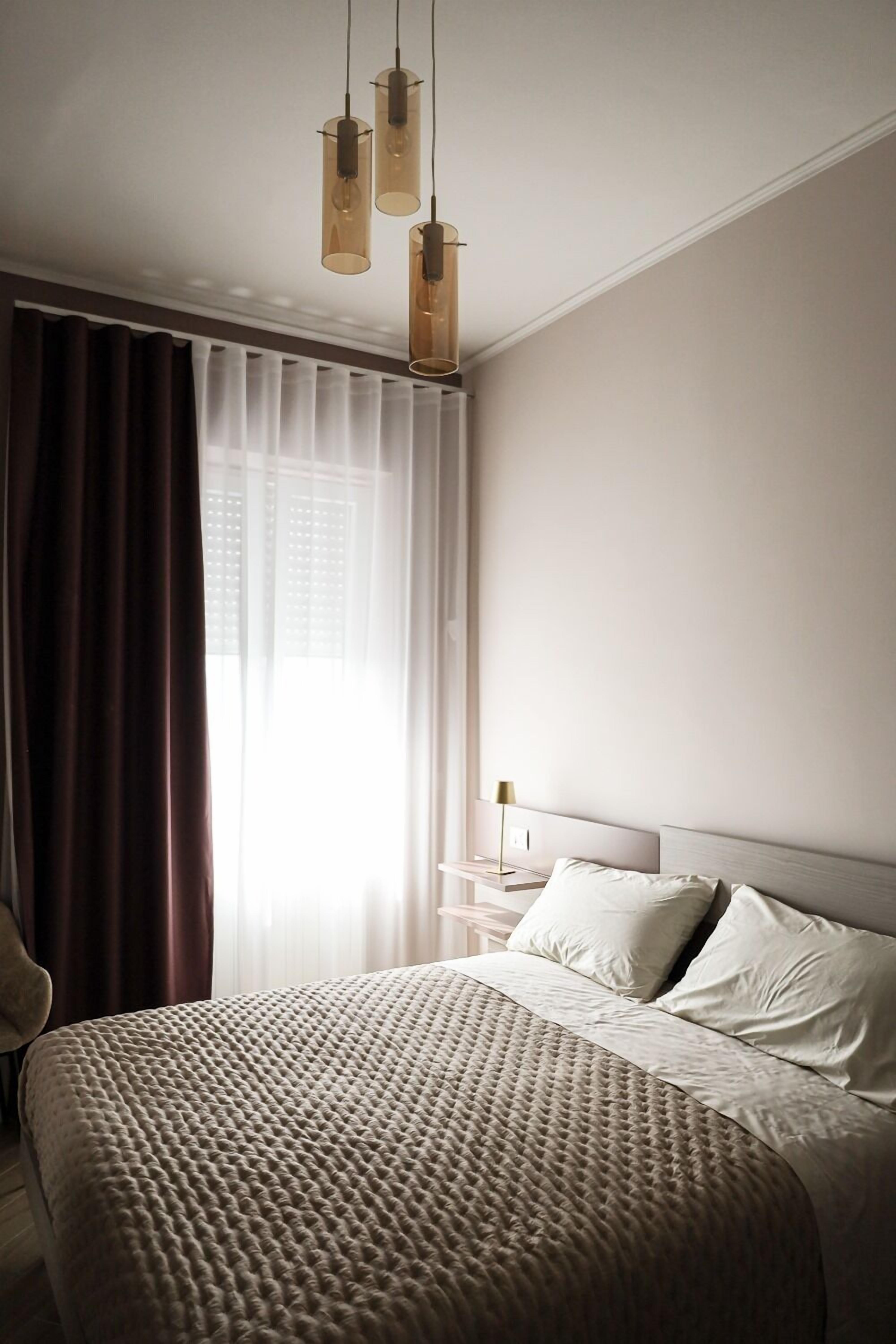 Un dormitorio con una cama grande, una mesita de noche, una lámpara y un ventanal con cortinas.