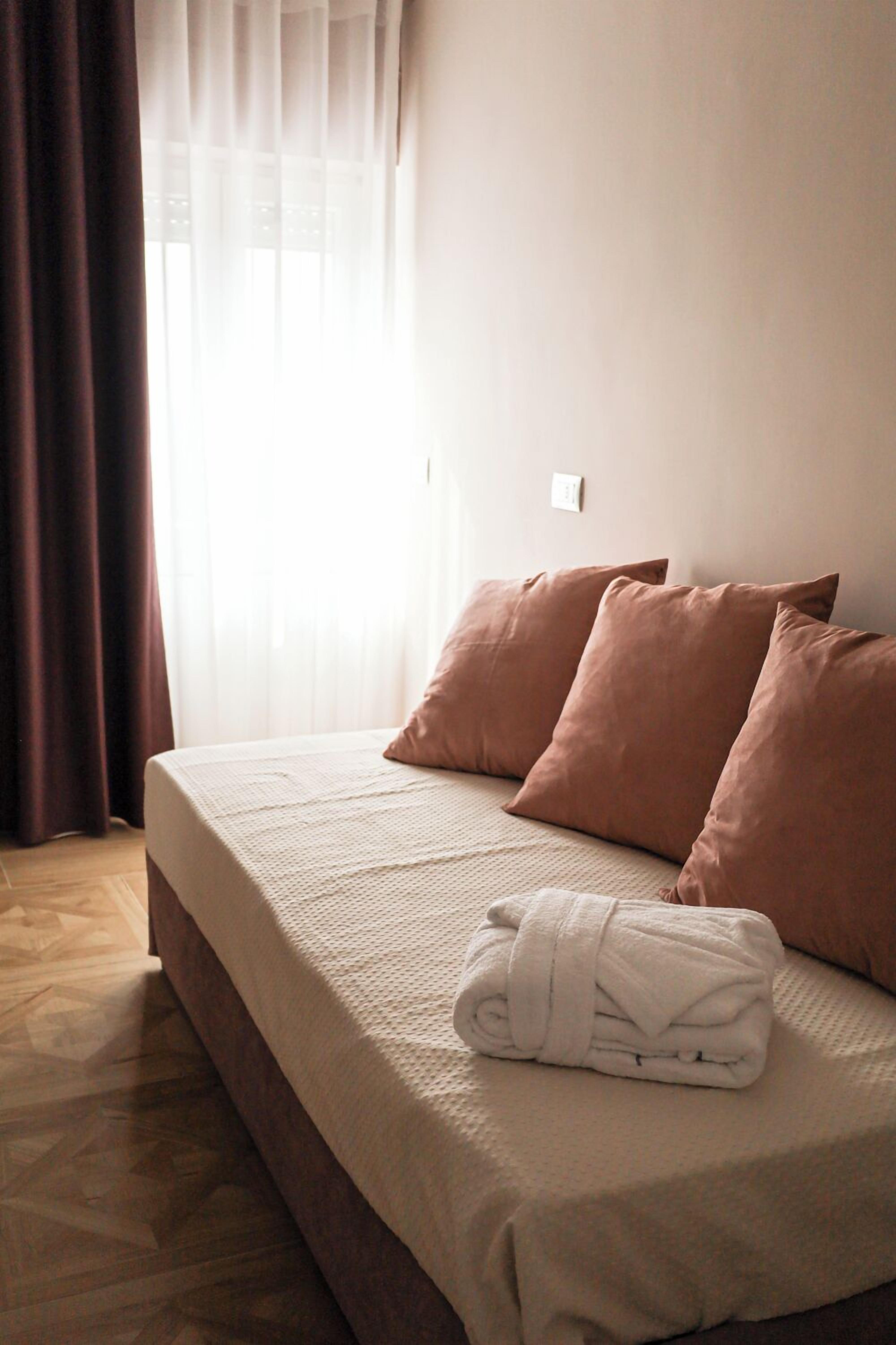 Una cama bien hecha con una manta blanca y dos almohadas marrones, una toalla blanca enrollada sobre la cama y una ventana con cortinas traslúcidas.