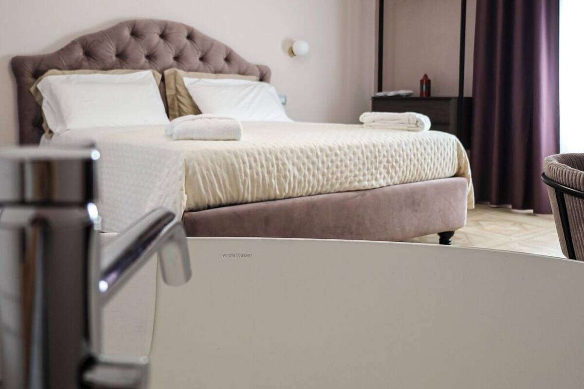 A bedroom with a tufted headboard, a bed with white and beige bedding, a bedside table, and a hanging coat rack.