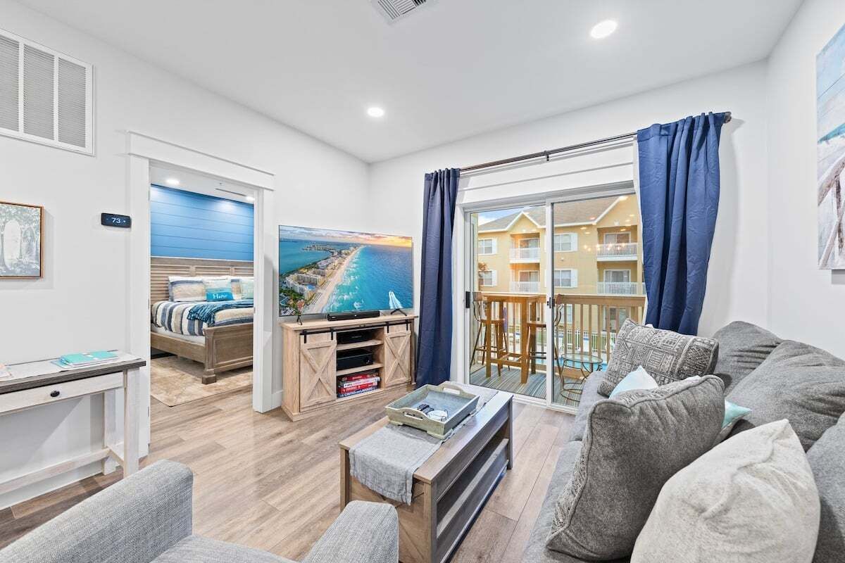 A modern living room with a sofa, a coffee table, and a TV stand. There is a painting of a coastal scene on the wall.