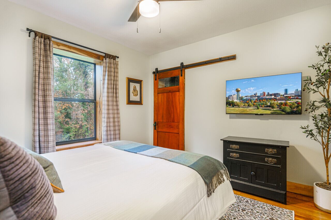 Un dormitorio con cama, cómoda, televisor, ventana con cortinas y una planta.