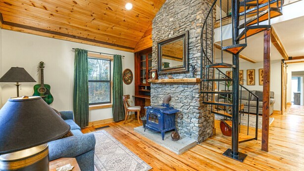 A cozy living room with a stone fireplace, a blue stove, a wooden ceiling, and a spiral staircase.