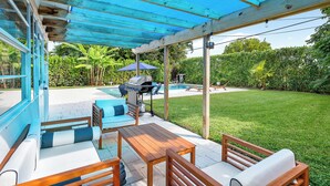 An outdoor patio with a wooden table and chairs, a barbecue grill, and a pool in the background.