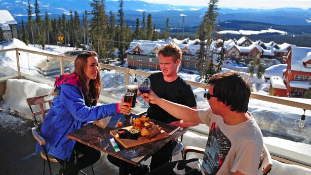 Three people toasting with drinks at a table with a view of snow-covered houses and mountains in the background. - Winter Escape On Perfection, Ski In/Out, Pool and Hot Tub (Big White)