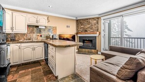 A kitchen with a stone backsplash, a fireplace, and a cozy seating area with a sofa and a small table. - Winter Escape On Perfection, Ski In/Out, Pool and Hot Tub (Big White)