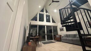 A spacious living area with a high ceiling, a black spiral staircase, and a large window providing natural light. - Modern Forest Chalet • Sleeps 6 • Starlink • Rivers & Caberfae • Pet-Friendly (Wellston)