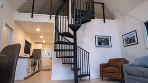 A spacious living area with a spiral staircase, a dining area, and a kitchen. - Modern Forest Chalet • Sleeps 6 • Starlink • Rivers & Caberfae • Pet-Friendly (Wellston)