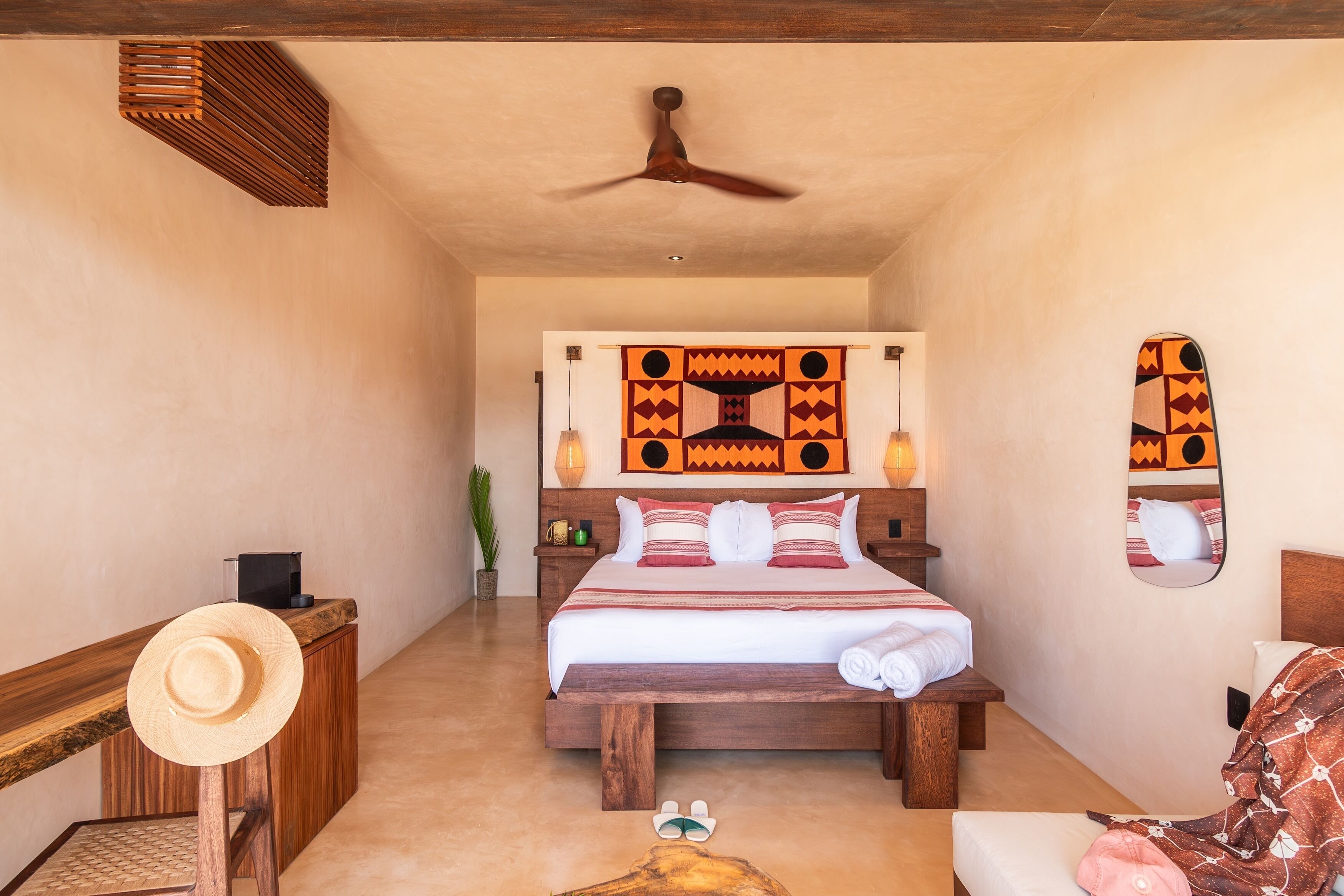 A bedroom with a wooden bed, a ceiling fan, a wall-mounted mirror, and a hat on a wooden stool.