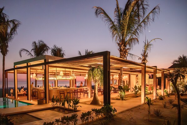 A modern outdoor bar area with a pool, surrounded by palm trees and plants, overlooking the ocean. - Casa Yati (Santa María Tonameca)
