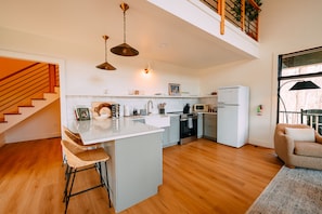 A modern kitchen with a central island, a dining area with bar stools, a staircase, and a view of the outdoors.