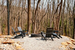 Ein Campingplatz mit vier Adirondack-Stühlen um ein Steinkaminfeuer, umgeben von Kies und Bäumen.