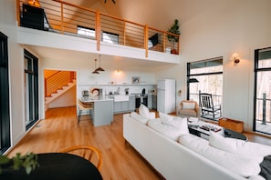 A spacious living area with a white sofa, wooden flooring, a kitchen with a central island, and a staircase leading to an upper level.