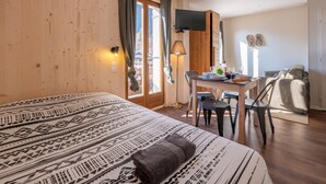 A hotel room with a large bed, a desk with chairs, a TV mounted on the wall, and a window with curtains. - Le Petit Aravis - panoramic balcony and village (La Giettaz)