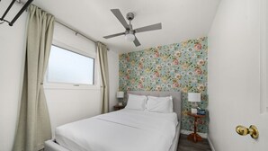 Une chambre à coucher bien rangée, avec un lit, un ventilateur de plafond, une fenêtre avec des rideaux, un papier peint à motifs floraux et une table de chevet avec une lampe.
