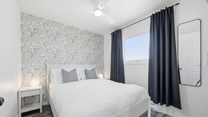 A bedroom with a white bed, a nightstand, a ceiling fan, a window with dark curtains, and a wall-mounted mirror.
