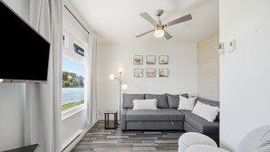A modern living room with a grey sofa, a flat-screen TV, a ceiling fan, and a view of a lake.