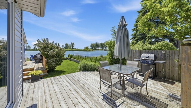 A wooden deck with a glass table and chairs, a barbecue grill, and a view of a lake. - Wellington Cottage at Twin Birch Suites (Prince Edward)