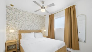 A bedroom with a bed, a nightstand, a ceiling fan, a window with curtains, and a mirror on the wall. - Wellington Cottage at Twin Birch Suites (Prince Edward)
