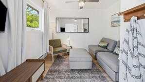 A modern room with a ceiling fan, a sofa, a chair, a small table, and a window with curtains. - Wellington Cottage at Twin Birch Suites (Prince Edward)